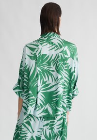 Camicia a maniche corte verde e azzurra con un design di foglie di palma, dal taglio rilassato e dalla texture liscia. La vista posteriore mostra il colletto e i dettagli.