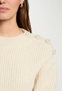 Cremfarbener Strickpullover mit geripptem Textur und dekorativen, strukturierten Knöpfen an der Schulter. Glatter, runder Halsauschnitt.