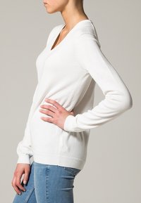 Witte langemouwen trui met een V-hals ontwerp, gemaakt van zachte stof, met ribgebreide manchetten en zoom, gedragen over blauwe jeans.