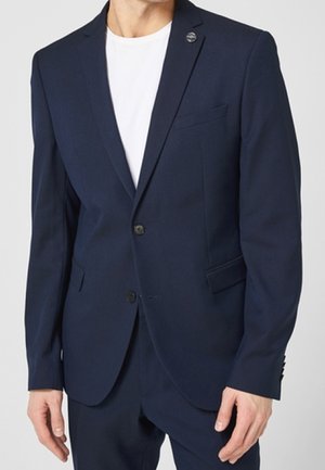 Man draagt een marineblauw colbert met twee knopen en een wit T-shirt met ronde hals eronder, bijgesneden ter hoogte van de nek en dijen.