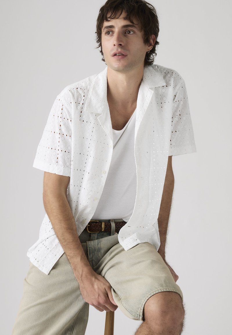 Junger Mann sitzt auf einem Hocker, trägt ein weißes, kurzärmeliges Hemd mit Lochmuster über einem weißen Tanktop und beige Shorts mit einem geflochtenen Gürtel.