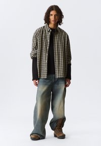 Chemise à carreaux beige et vert foncé sur un top noir à manches longues, associée à un jean bleu évasé et des bottes beige.