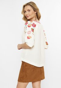 Bluză albă cu broderie florală colorată pe umeri, mâneci scurte și o croială lejeră, asortată cu o fustă din Suede maro.