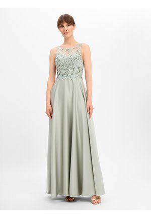 Frau trägt ein ärmelloses, blassgrünes Kleid mit floraler Spitzen-Bodice und fließendem Rock, steht vor einem schlichten weißen Hintergrund.