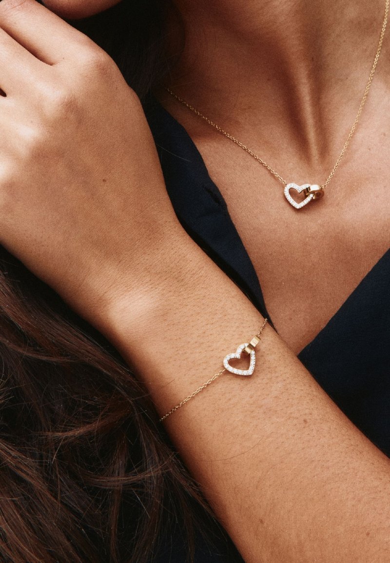 Zelta kaklarota un rokassprādze ar sirds formas kuloniem, ko nēsā sieviete ar tumšu apģērbu un brūnu ādu.