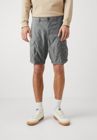 Pantalones cortos cargo grises con bolsillos laterales, cinturilla elástica y cierre de botón. Llevado con calcetines blancos y zapatillas sobre un fondo neutro.