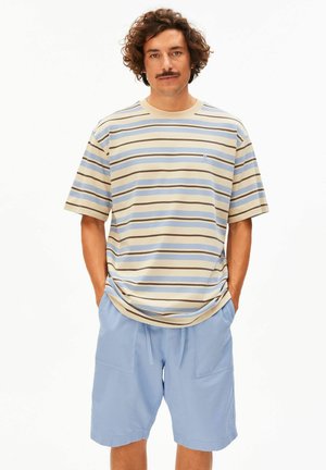 Man met krullend haar en snor die een beige, blauw en bruin gestreept T-shirt en lichtblauwe korte broek draagt met handen in de zakken.