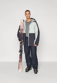 Skibekleidung mit einer zweifarbigen Kapuzenjacke in Hellgrau und Marineblau, kombiniert mit marineblauen Hosen. Das Modell hält ein Snowboard.