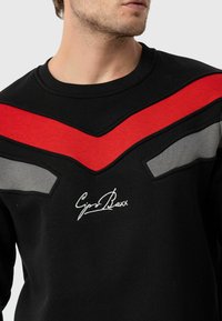 Zwarte sweatshirt met ronde hals, voorzien van rode en grijze chevronsaccenten en geborduurd logo op de borst. Soepele textuur.