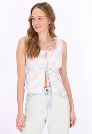 Fiatal nő hosszú hajjal, ujjatlan rózsaszín-fehér foltmintás felsőt, világos színű farmernadrágot és rózsaszín gyűrűt visel az ujján.