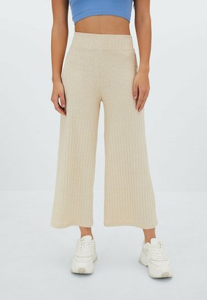 Persona con pantalones anchos color beige con textura acanalada, zapatillas blancas gruesas y un top corto azul sobre un fondo blanco liso.