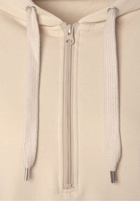 Sweat à capuche beige avec fermeture éclair, présentant une texture de tissu lisse, des cordons de serrage texturés contrastants, et une fermeture éclair métallique avec un tirette circulaire.