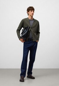 Man som bär en olivgrön kofta, grå t-shirt, blå jeans och bruna skor, håller en svart läderportfölj mot en ljus, enfärgad bakgrund.