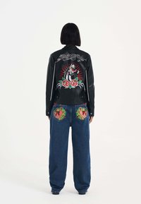 Svart läderjacka med broderad design av en krabba och rosor på ryggen. Blå denimbyxor har färgglada mönstrade fickor.