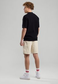 Polo nero a righe con maniche corte, pantaloni corti crema, sneakers bianche, calze a righe rosse e bianche, vista da dietro, sfondo neutro.
