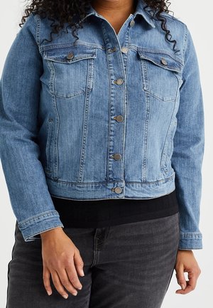 Kvinde iført en blå denimjakke med metal knapper og klaplommer på brystet over en sort top og mørke jeans.