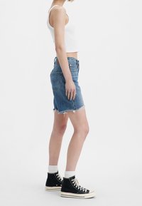 Jupe en denim bleu clair, ourlet effiloché, longueur au milieu de la cuisse, associée à des baskets montantes noires et des chaussettes blanches à la cheville.