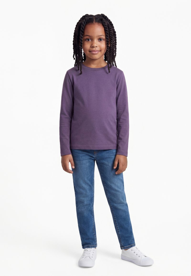Jeune fille aux cheveux torsadés portant un t-shirt à manches longues violet, un jean bleu et des baskets blanches, debout devant un fond blanc.