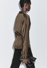 Brown satin blouse with a relaxed fit, gathered waist, and long sleeves. Paired with black pants and a small black handbag.
