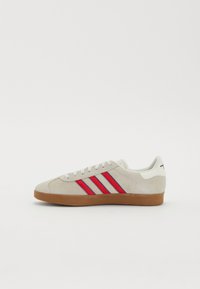 Sneakers din suede gri deschis cu accente roșii și albastre. Au o talpă plată din cauciuc și un design clasic cu șireturi. Pe lateral au trei dungi orizontale.