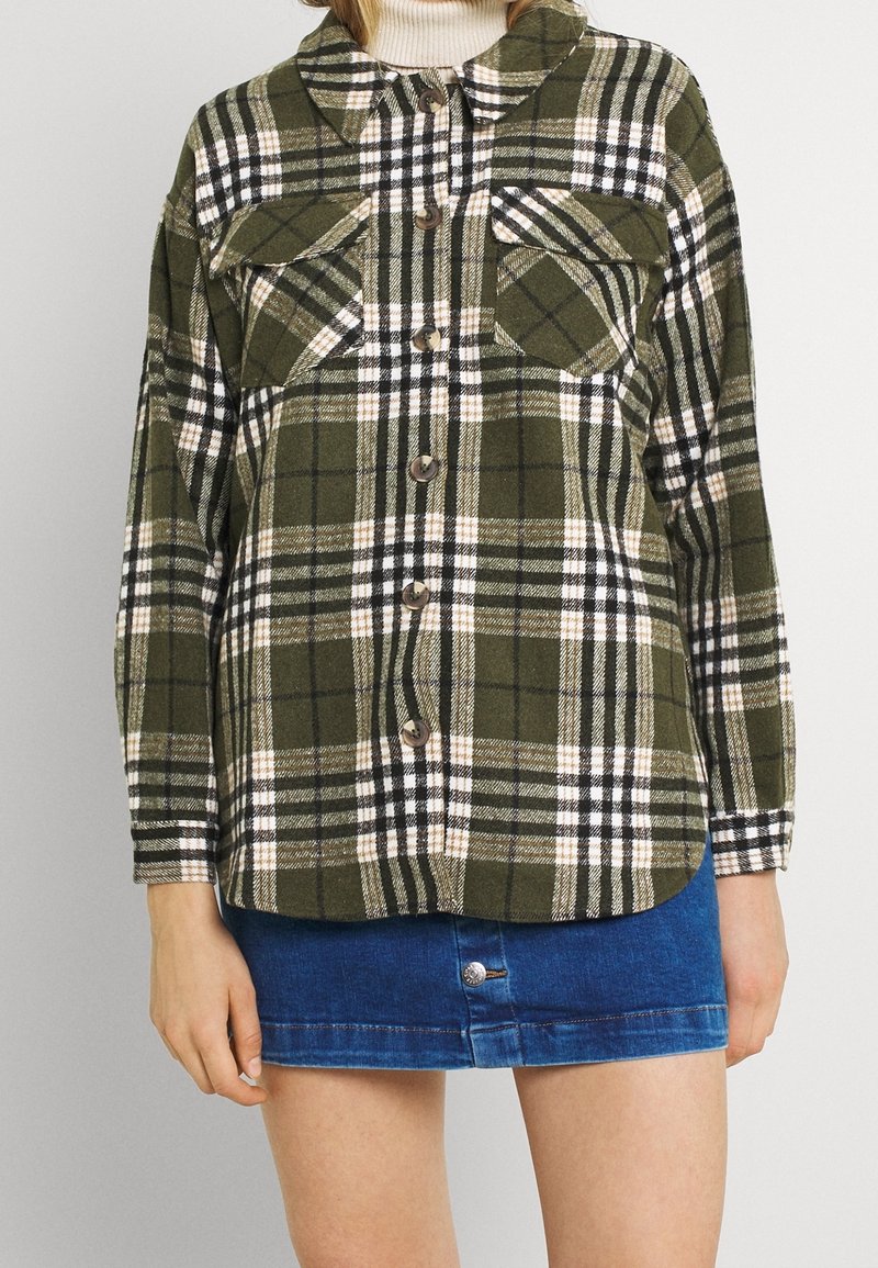 Chemise à boutons à carreaux verts, noirs et blancs, avec deux poches poitrine et un ourlet arrondi. Confectionnée dans un tissu texturé.