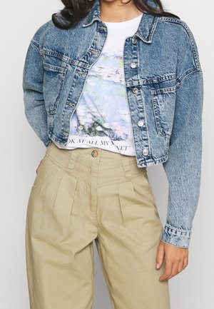 Person iført cropped blå denimjakke over hvid grafisk t-shirt og højtaljede beige plisserede bukser, stående med den ene hånd ned langs siden.
