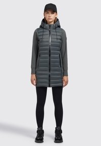 Padded šedá bunda s kapucí, zipem v přední části a bočními kapsami. V kombinaci s černými legínami a botami s elegantním, moderním designem.