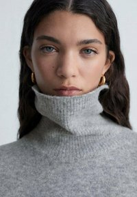 Pull à col roulé gris à côtes avec un col haut, une texture douce et un motif subtil de tâches, associé à des boucles d'oreilles rondes en or.