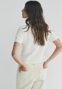Hvid kortærmet strikket sweater med tekstureret mønster og ribbet kant, kombineret med lys beige bukser, hænderne i lommerne, set bagfra.