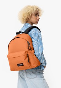 Zaino arancione in nylon con spallacci neri, tasca frontale e cerniere; design minimalista su una camicia azzurro chiaro a stampa.