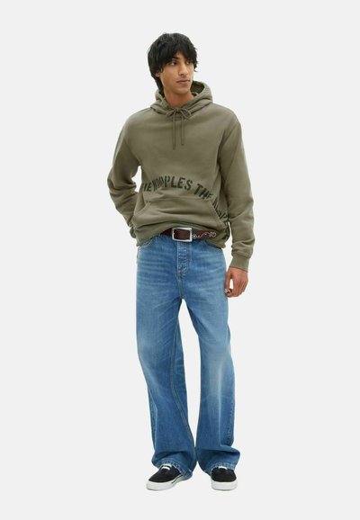 Haut à capuche vert olive avec poches latérales et texte en gras, associé à un jean flare bleu clair et des baskets noires. Tenue décontractée, design simple.