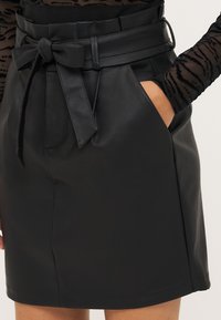 Μαύρη φούστα από συνθετική δερματίνη με ζώνη που διαθέτει λεπτομέρεια κόμπο, πλαϊνές τσέπες και λεία υφή, ραμμένη σε ίσια γραμμή.