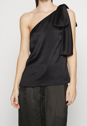 Kobieta w czarnej satynowej bluzce na jedno ramię z dużą kokardą na ramieniu, w połączeniu z luźnymi czarnymi spodniami skróconymi do połowy tułowia.