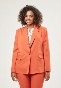 Oranger Blazer mit einem einzigen goldenen Knopf, Reverskragen und zwei Vorder-taschen, kombiniert mit einer floral gemusterten Bluse. Glatte Textur, taillierte Passform.