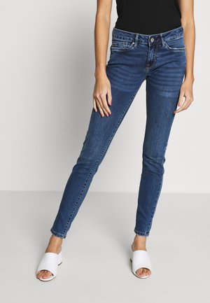 Kobieta w ciemnoniebieskich obcisłych dżinsach i białych sandałach na obcasie z odkrytymi palcami stoi na tle jednolitego jasnego tła.