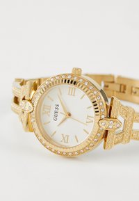 Montre-bracelet dorée avec un cadran blanc, des chiffres romains et une lunette ornée de cristaux. Dispose d'un design de bracelet décoratif doré.