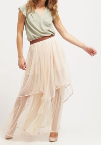 Lys grøn bluse med flæseærmer, parret med en flydende, gennemsigtig beige plisseret nederdel med asymmetrisk kant. Neutrale pumps.