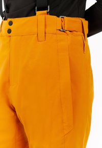 Oranje waterdichte broek met een gladde textuur, verstelbare taille, twee zwarte knopen, zijzakken en versterkte stiksels.