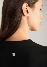 Czarna bluzka z okrągłym dekoltem oraz małym metalowym logiem na plecach, wyróżniająca się gładką teksturą i minimalistycznym designem.