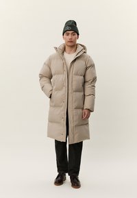 Άνδρας που φοράει μακρύ μπεζ παλτό με επένδυση, μαύρο σκουφάκι, μαύρο παντελόνι και σκούρα καφέ παπούτσια, στέκεται μπροστά σε απλό ανοιχτό φόντο.