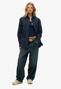 Veste boutonnée marine portée sur un débardeur marine, associée à un jean large bleu foncé et des baskets beiges, avec une ceinture décorative.