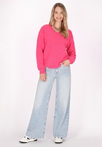 Pullover fuzzy di un brillante rosa con scollo a V e maniche lunghe, abbinato a jeans larghi azzurri e scarpe bianche con accenti neri.