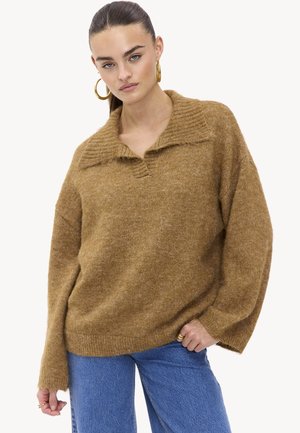 Bruine, oversized gebreide trui met een gestructureerde kraag en lange mouwen, gecombineerd met lichtblauwe spijkerbroeken. Het materiaal lijkt zacht en comfortabel.