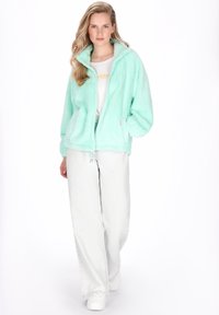 Menta zöld polárdzseki cipzárral elöl és magas gallérral, világosszürke, bő szabású farmerrel és fehér tornacipővel párosítva.