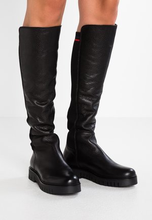 Sorte knæhøje støvler med kraftige såler båret på bare ben, med tekstureret læder foran og strækbare ribstrikkede paneler bagpå.