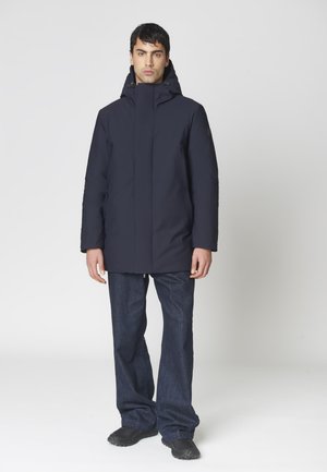 Hombre con un abrigo de invierno con capucha de color azul marino oscuro, vaqueros azul oscuro y zapatillas negras, de pie contra un fondo claro y liso.