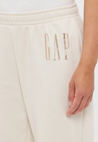 Pantaloni della tuta color crema con una texture morbida, caratterizzati da un logo "GAP" metallic oro sulla coscia sinistra e una cintura elastica.