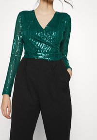 Haut en sequins vert avec un col en V ; corsage ajusté et manches longues ; associé à un pantalon noir taille haute avec poches latérales.