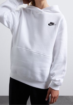 Person iført en hvid Nike-hoodie med en frontlomme, sort Nike-logo på brystet og sorte bukser mod en ensartet lys baggrund.