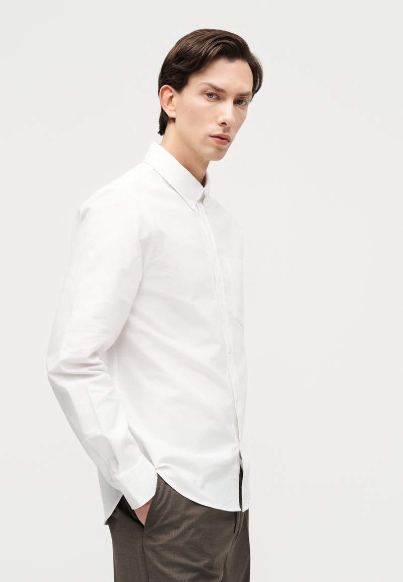 Junger Mann mit dunklem Haar, der ein weißes Hemd mit Knöpfen und graue Hose trägt, steht mit den Händen in den Taschen vor einem einfarbigen Hintergrund.